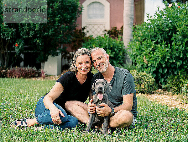 hispanic couple sits in their front yard  smiling with their puppy