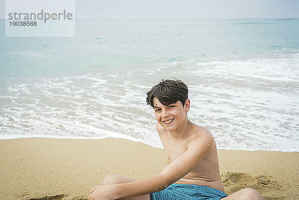 Lächelnder kleiner Junge in Badebekleidung sitzt am Strand und blickt in die Kamera