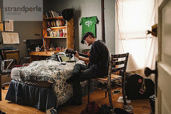 Kaukasischer Teenager spielt Gitarre  während er im Stuhl in einem unordentlichen Schlafzimmer sitzt