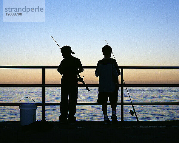 Rückansicht von zwei Jungen mit Angelruten  die in der Dämmerung auf einem Pier aufragen. Kalifornien.