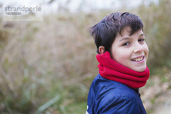 Rückansicht eines lächelnden Teenagers  der einen roten Schal trägt  während er draußen in der Natur in die Kamera blickt
