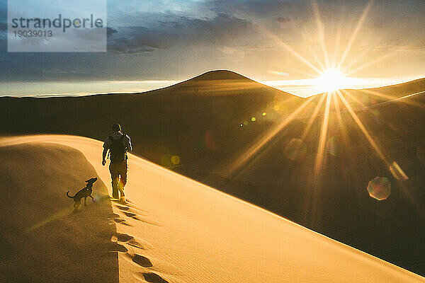 Wanderer und sein Hund klettern an einem windigen Abend bei Sonnenuntergang auf eine Sanddüne
