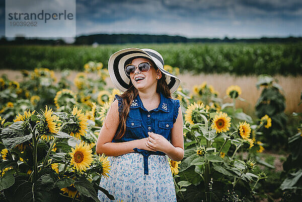 Mädchen mit Sonnenbrille und Strohhut lacht in einem Sonnenblumenfeld