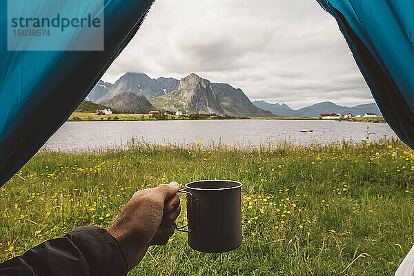 Die Hand eines Mannes hält einen Becher in einem Campingzelt mit Seeblick in Norwegen