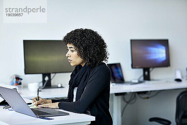 Seitenansicht einer selbstbewussten Geschäftsfrau  die auf einen Klebezettel schreibt  während sie im Büro ihren Laptop an der Wand benutzt