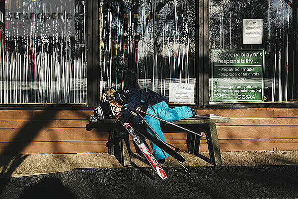 Little girl wearing snow gear rests on bench at ski shop after cross country skiing in North Dakota