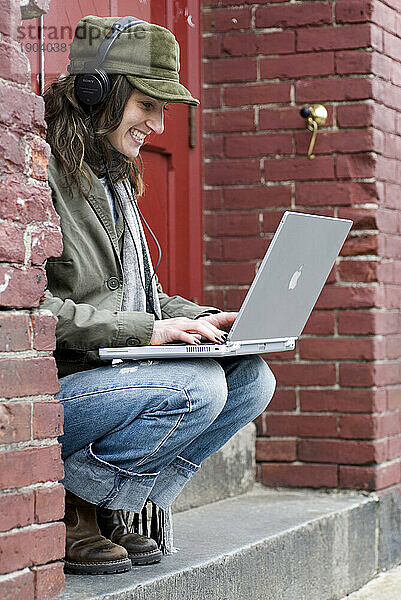 Hippe Frau entspannt sich vor roter Tür  während sie am Laptop arbeitet und Musik hört.