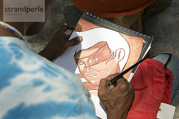 Ein junger männlicher kubanischer Straßenkünstler malt auf einem Skizzenblock ein Aquarell des Gesichts eines Mädchens. Centro Havana  La Habana  Kuba