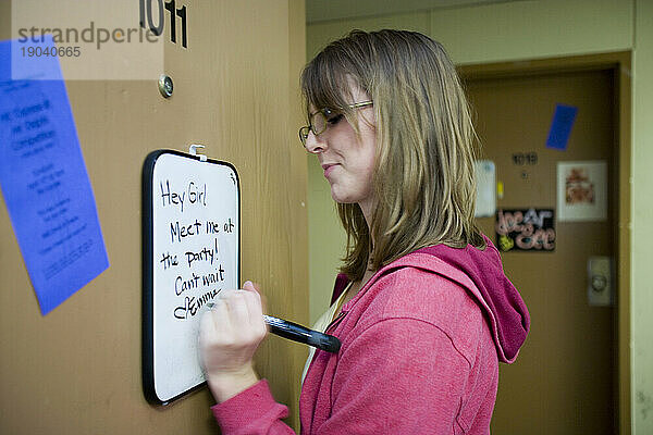 Eine junge kaukasische Studentin schreibt eine Notiz an ihre Freunde an die weiße Tafel an ihrer Wohnheimtür.