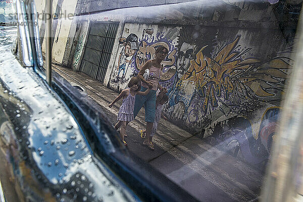 Eine Frau geht mit zwei kleinen Kindern spazieren  gesehen im Spiegelbild eines Oldtimerfensters. Alt-Havanna oder La Habana Veija  La Habana  Kuba