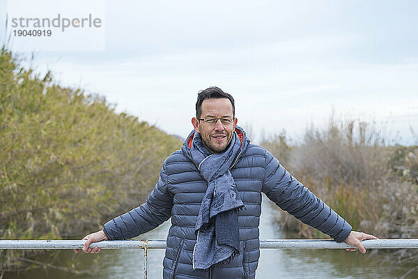 Ganzkörperporträt eines lächelnden erwachsenen Mannes  der am Geländer einer Brücke steht und tagsüber mit der Kamera auf den Fluss blickt
