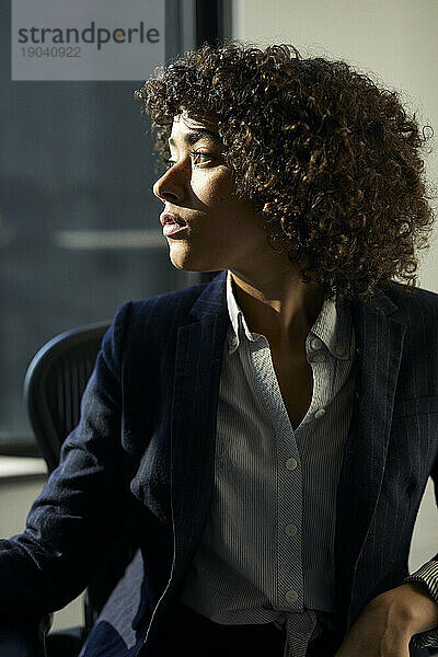 Nahaufnahme einer nachdenklichen Geschäftsfrau mit lockigem Haar  die wegschaut  während sie im Büro sitzt