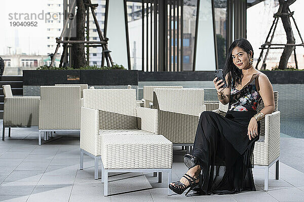 schöne thailändische Frau  die in die Kamera schaut  während sie ihr Smartphone benutzt