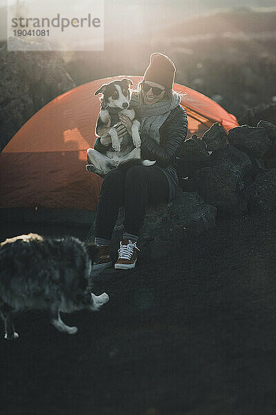 Glückliche Camperin hält ihren Hund bei Sonnenuntergang mit orangefarbenem Zelt