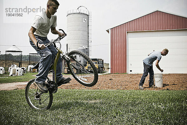 Ein Mann bereitet den Boden für den Pflanzenanbau im Hinterhof des Bauernhauses vor  während im Vordergrund ein Junge mit dem Fahrrad fährt.