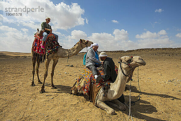Einem Mann mittleren Alters wird auf ein Kamel geholfen  während sein Freund zusieht