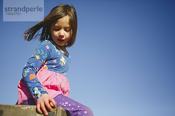 Ein kleines Mädchen sitzt friedlich auf einer Kiste vor dem blauen Himmel und dem Wind im Haar