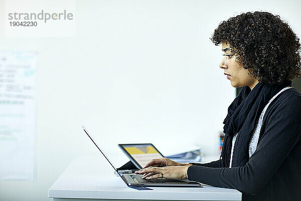 Seitenansicht einer seriösen Geschäftsfrau  die einen Laptop-Computer auf dem Schreibtisch benutzt  während sie im Büro an der Wand steht