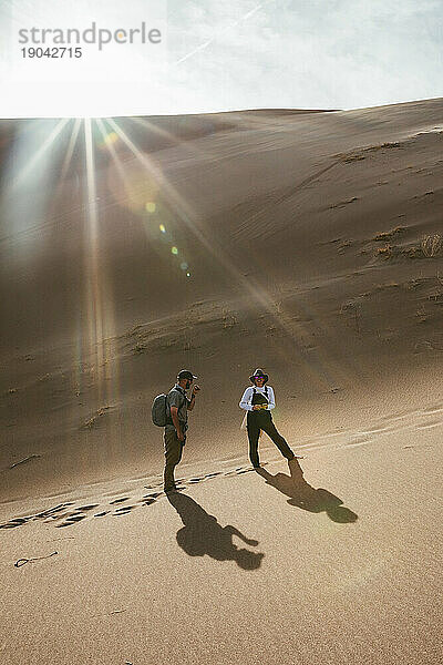 Zwei Wanderer in Sanddünen bleiben stehen und lächeln im Sonnenlicht