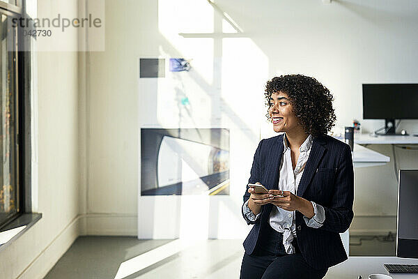 Lächelnde Geschäftsfrau mit Smartphone schaut weg  während sie im Büro an der Wand steht