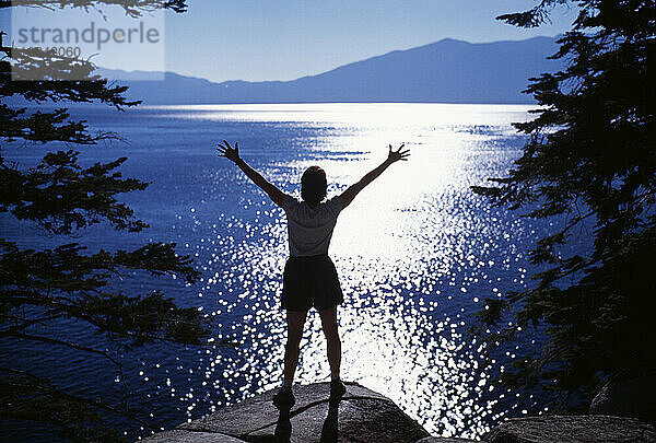 Eine Frau im Gegenlicht mit ausgestreckten Armen steht auf einem Felsen mit Blick auf Lake Tahoe  Kalifornien.