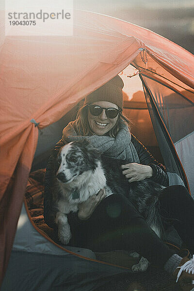 Lächelnde Camperin und ihr Hund entspannen sich bei Sonnenuntergang im orangefarbenen Zelt
