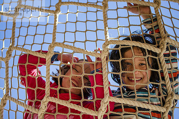 two laughing children peer down through a rope net against blue sky