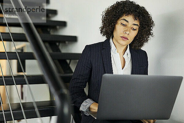 Selbstbewusste  ernsthafte Geschäftsfrau mit lockigem Haar benutzt Laptop  während sie auf Stufen im Büro sitzt