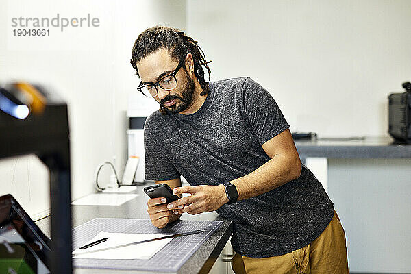 Selbstbewusster Geschäftsmann benutzt Smartphone  während er im Büro am Tisch steht