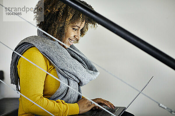 Seitenansicht einer lächelnden Geschäftsfrau mit lockigem Haar  die einen Laptop benutzt  während sie auf einer Treppe im Büro sitzt