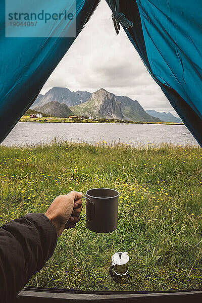 Man's hand holding a mug in a camping tent with a lake view in Norway