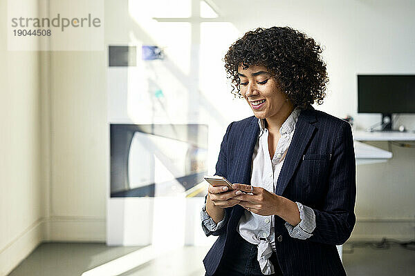 Lächelnde Geschäftsfrau benutzt Smartphone  während sie im Büro an der Wand steht