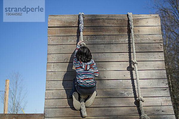 Ein kräftiges Kind zieht sich draußen an einer Holzwand einen Seilgarten hinauf