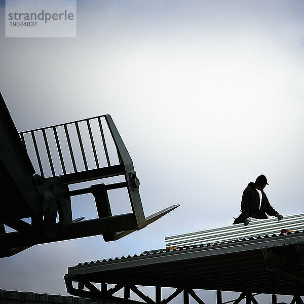 Silhouette eines Gabelstaplers und eines Bauarbeiters auf einem Dach vor bewölktem Himmel.