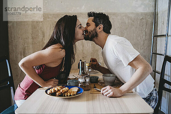 Junges glückliches Paar küsst sich beim Frühstück am Küchentisch