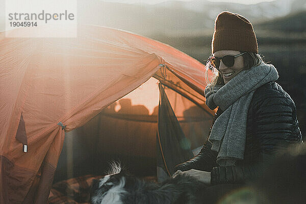 Fröhliche Camperin mit Mütze bei Sonnenuntergang vor dem orangefarbenen Zelt