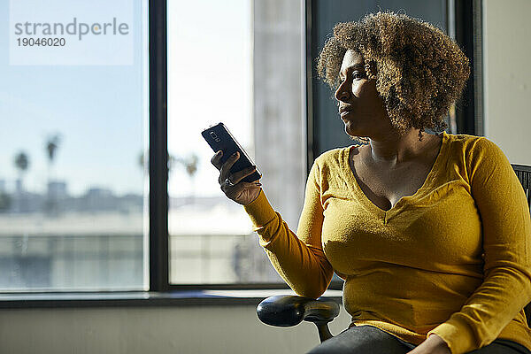 Seriöse Geschäftsfrau benutzt Mobiltelefon  während sie im Büro auf einem Stuhl am Fenster sitzt