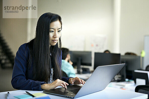 Selbstbewusste Geschäftsfrau benutzt Laptop-Computer auf dem Schreibtisch  während sie im Büro an der Wand steht
