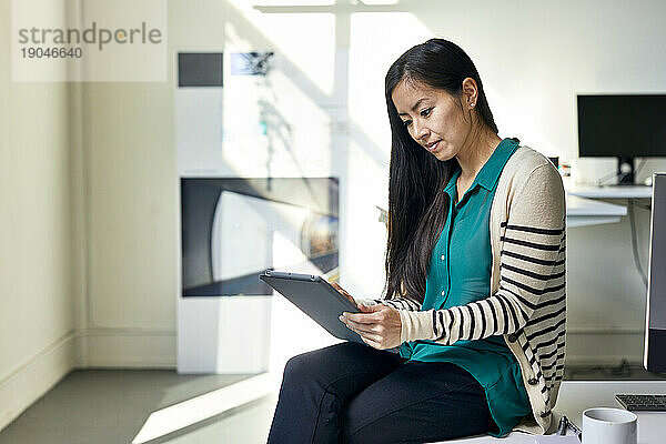 Selbstbewusste Geschäftsfrau nutzt Tablet-Computer  während sie im Büro auf dem Schreibtisch an der Wand sitzt