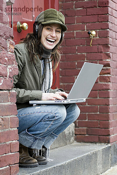 Hippe Frau entspannt sich vor roter Tür  während sie am Laptop arbeitet und Musik hört.