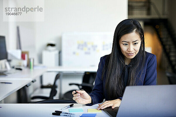 Selbstbewusste Geschäftsfrau schreibt auf Klebezetteln  während sie im Büro ihren Laptop an der Wand hält