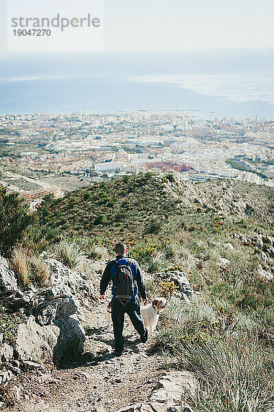 Mann wandert mit Hund einen Hügel hinunter und blickt auf die Stadt und das Meer in Malaga