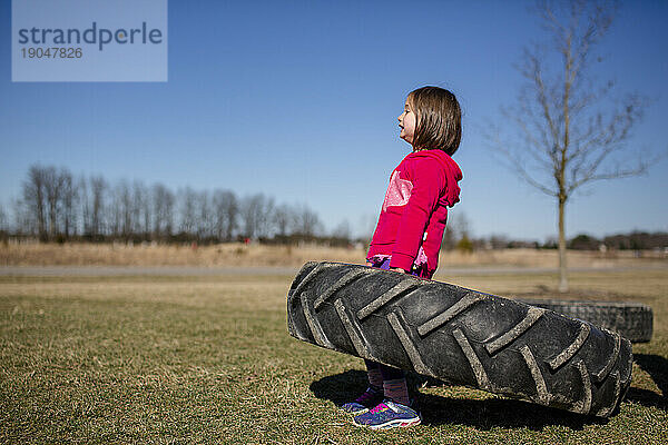 Ein starkes kleines Mädchen kämpft darum  einen großen Reifen in einem Park hochzuhalten