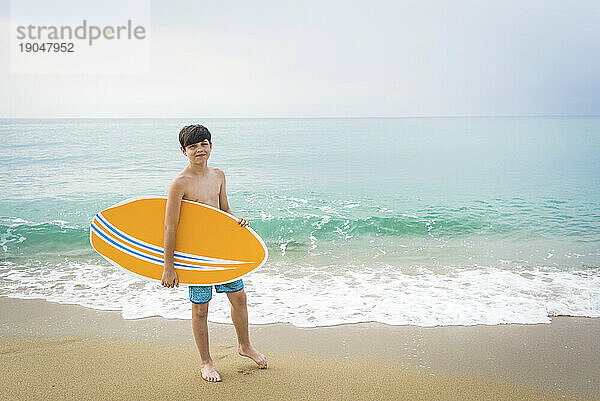 Porträt eines fröhlich lächelnden Teenagers mit gelbem Surfbrett  der am Strand steht