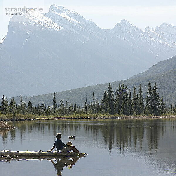 Mann sitzt am Ende eines Baumstamms im Alpensee  dahinter Mount Rundle