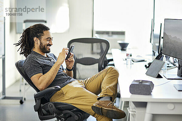 Seitenansicht eines lächelnden Geschäftsmannes mit Dreadlocks  der sein Mobiltelefon benutzt  während er auf einem Stuhl im Büro sitzt