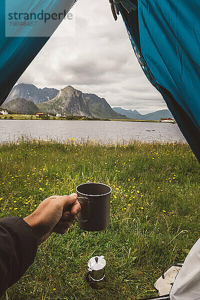 Die Hand eines Mannes hält einen Becher in einem Campingzelt mit Seeblick in Norwegen