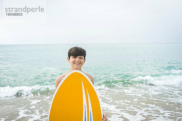 Vorderansicht eines lächelnden jungen Teenagers  der am Strand steht  ein Surfbrett in der Hand hält und in die Kamera blickt