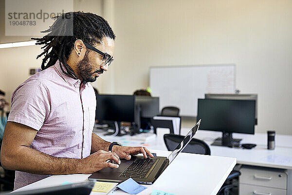 Seitenansicht eines Geschäftsmannes mit Dreadlocks  der einen Laptop auf dem Schreibtisch im Büro benutzt