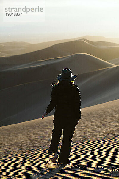 Frau mit Filzhut wandert bei Sonnenuntergang durch die Wüstendünen